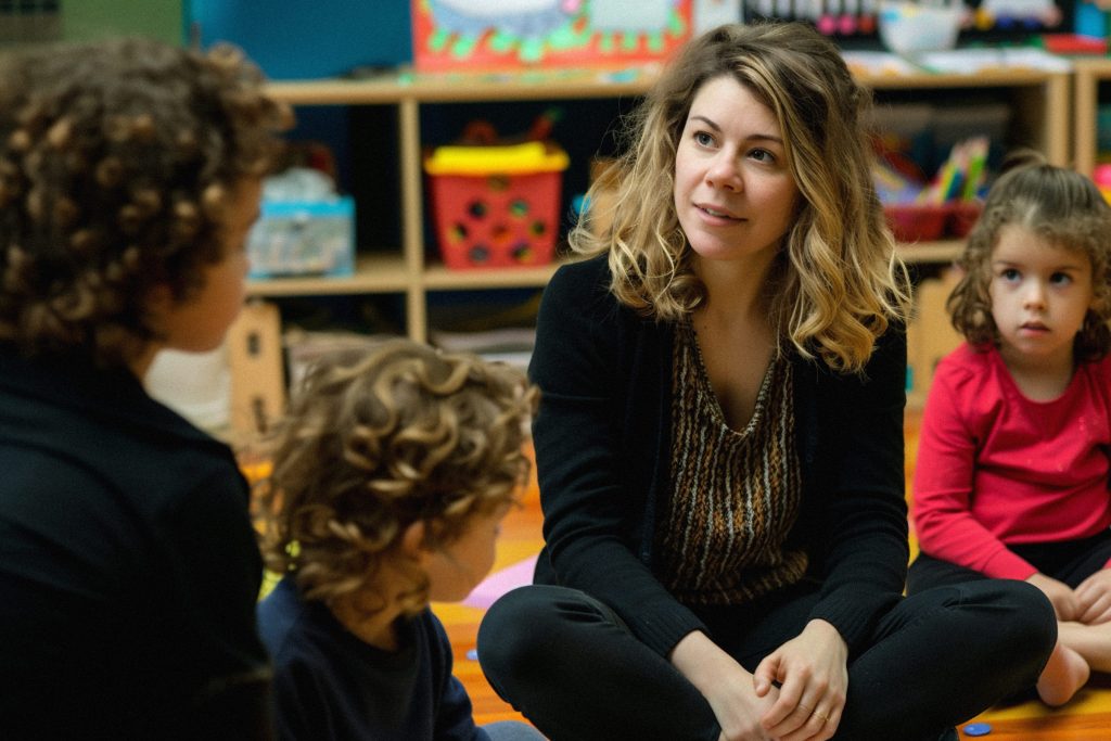 Eine Erzieherin sitzt im Schneidersitz gemeinsam mit Kindern auf dem Boden in einem kindgerecht gestalteten Raum. Kontext: Job-Angebot Erzieherin, Erzieher, Sozialpädagogische Assistentin, Sozialpädagogischer Assistent, Erziehungshelferin, Erziehungshelfer