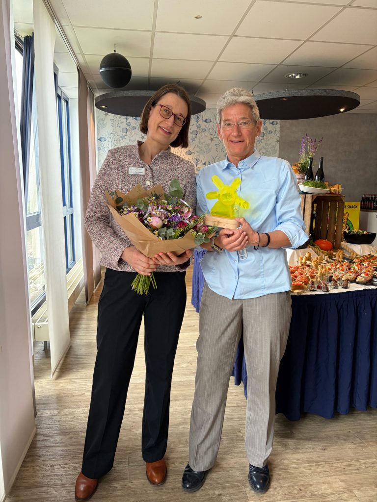 Zwei Frauen lächeln in einem hellen Raum bei einer feierlichen Übergabe in der Ostseeklinik Grömitz. Links steht die neue Klinikleiterin Frau Reeker-Meyer in einem grau melierten Blazer und hält einen bunten Blumenstrauß. Rechts daneben hält die scheidende Klinikleiterin Frau Löwner, gekleidet in einem hellblauen Hemd, eine gelbe Trophäe in Form einer Windmühle. Im Hintergrund ist ein Buffet mit Fingerfood und herbstlicher Dekoration aufgebaut.