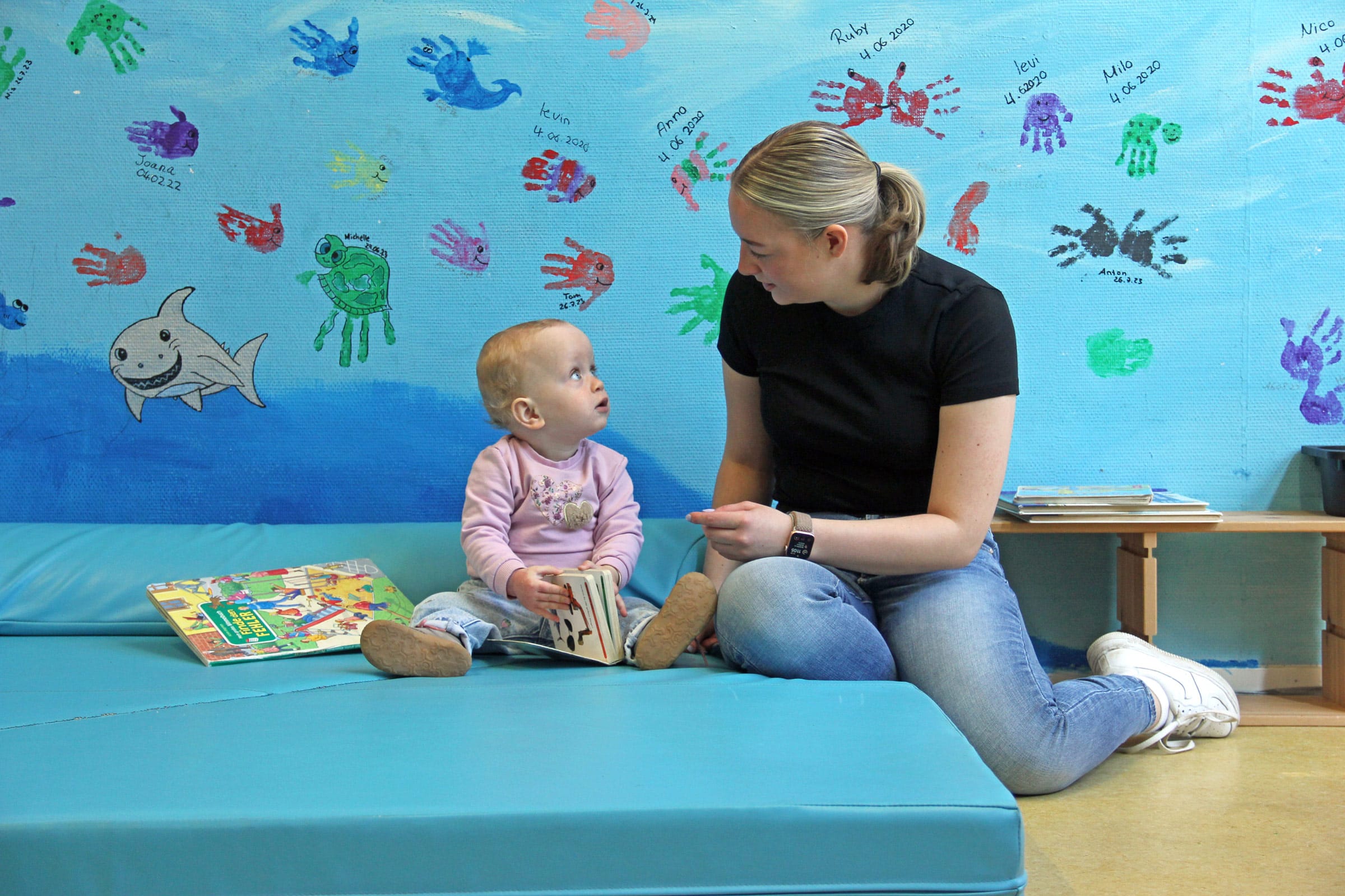 Eine Mutter und ein Kleinkind sitzen gemeinsam auf einer blauen Spielmatte in der Ostseeklinik Grömitz. Das Kind hält ein Bilderbuch in den Händen und schaut die Mutter aufmerksam an, während diese sich ihm freundlich zuwendet. Im Hintergrund ist eine hellblaue Wand mit bunten Handabdruck-Malereien und Namen zu sehen.