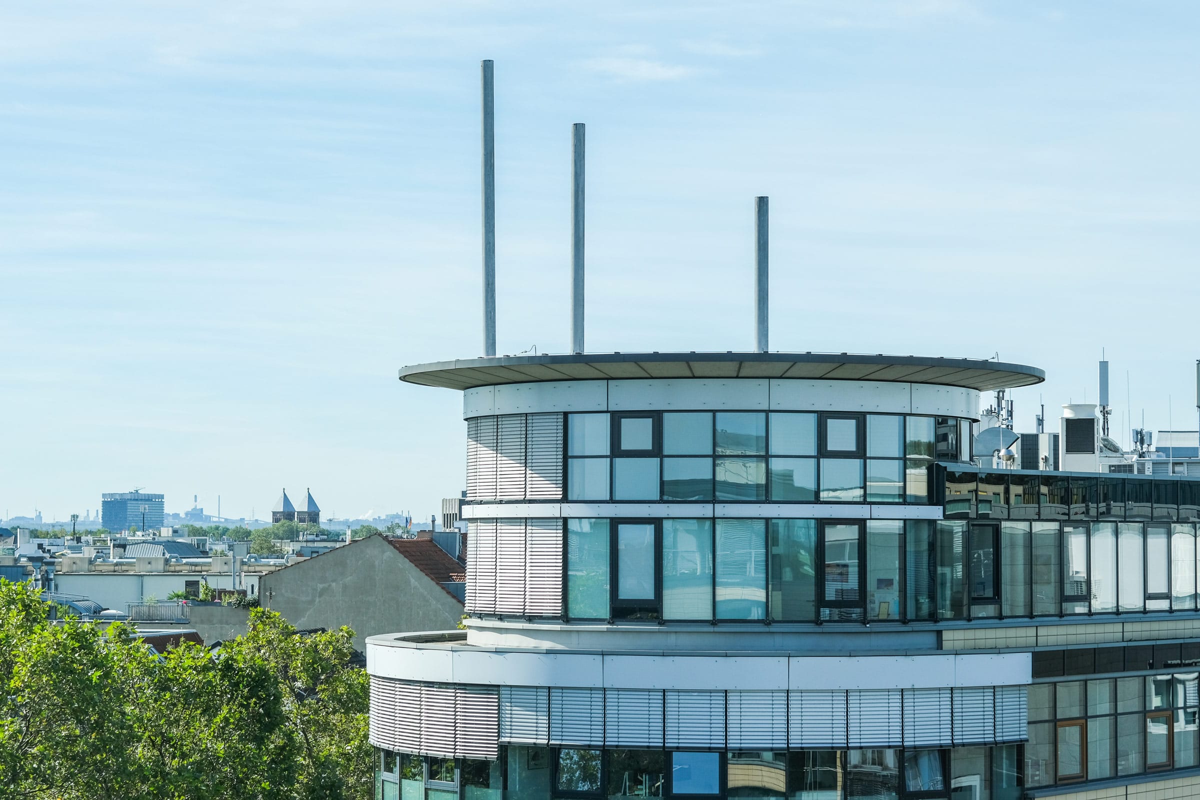 Nahaufnahme der oberen Stockwerke eines modernen, zylinderförmigen Glasgebäudes mit drei markanten, vertikalen Metallantennen auf dem Dach vor einem hellblauen Himmel. Im Hintergrund sind die Spitzen einer Kathedrale und die Dächer einer Stadt erkennbar.