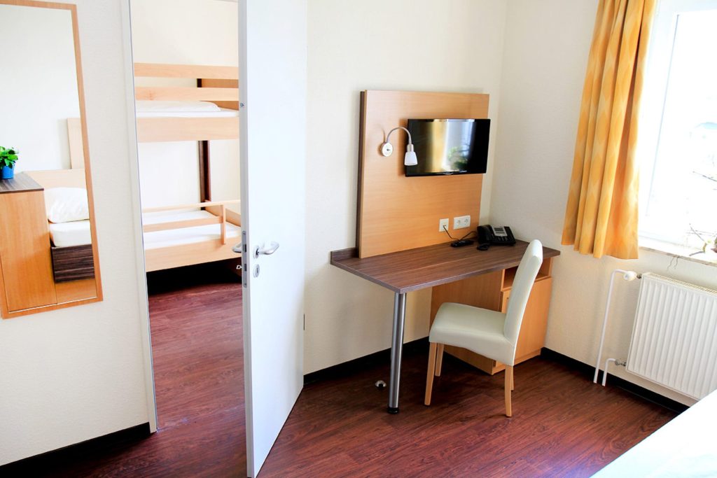 Helles, funktionales Patientenzimmer mit Blick durch eine offene weiße Tür in einen angrenzenden Schlafraum mit Holz-Etagenbetten. Im Vordergrund steht ein dunkler Schreibtisch mit einem weißen Stuhl, darüber ist ein Flachbildfernseher an einem Holzpaneel montiert. Ein Fenster mit gelben Vorhängen lässt Tageslicht in den Raum mit dunklem Laminatboden.