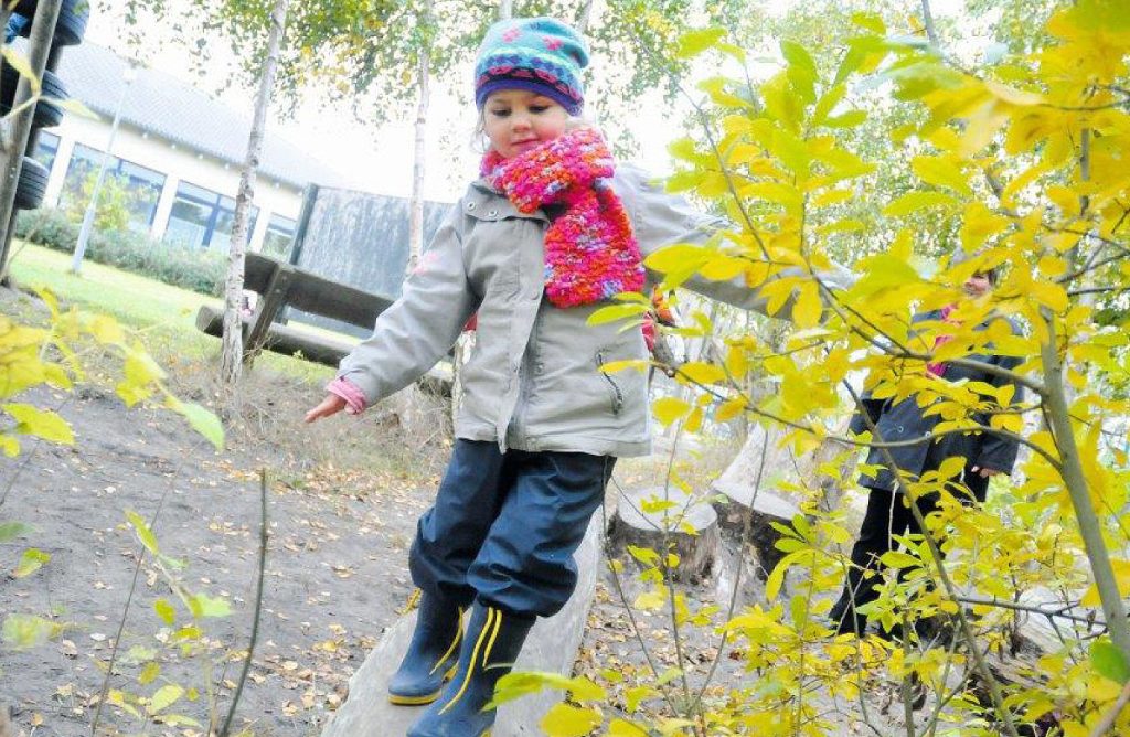 Ein Kind in Winterkleidung balanciert auf einem Holzstamm auf dem Außengelände 'Grömelland' der Ostseeklinik Grömitz, umgeben von herbstlich gelben Blättern.