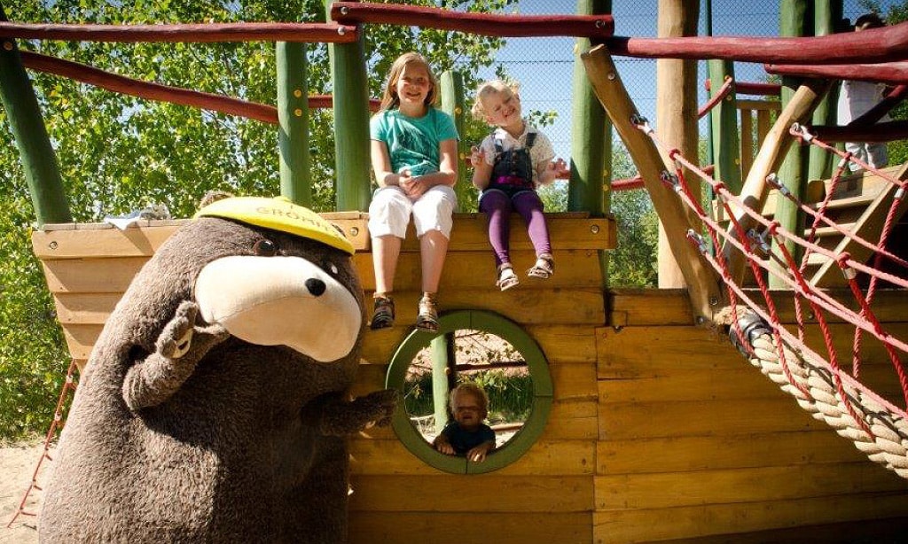 Zwei Kinder sitzen auf einem hölzernen Spielschiff im Außenbereich der Ostseeklinik Grömitz, während das Klinikmaskottchen Grömel, ein großer brauner Bär mit gelber Kappe, daneben steht und winkt.