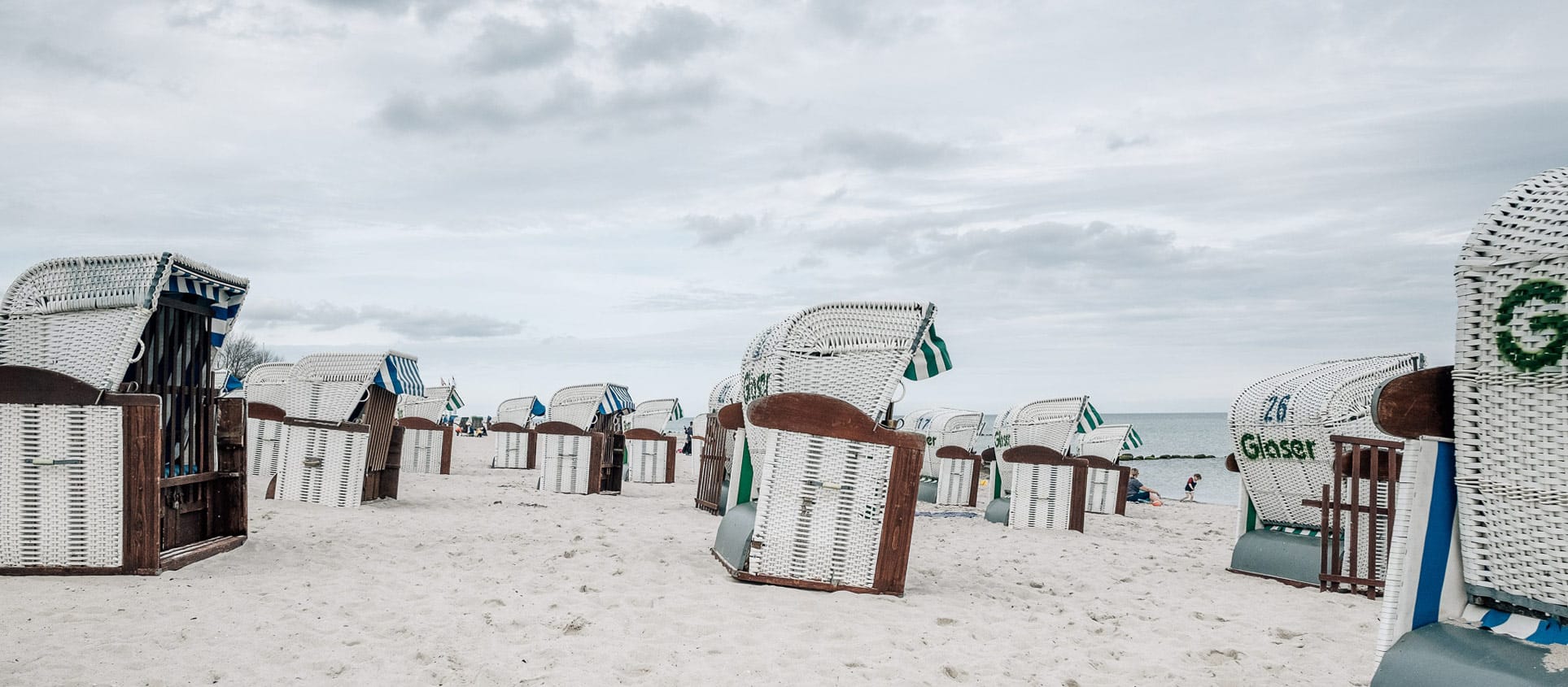 Zahlreiche weiß-blaue Strandkörbe stehen im hellen Sand am Strand an der Ostseeklinik Grömitz unter einem bewölkten Himmel.