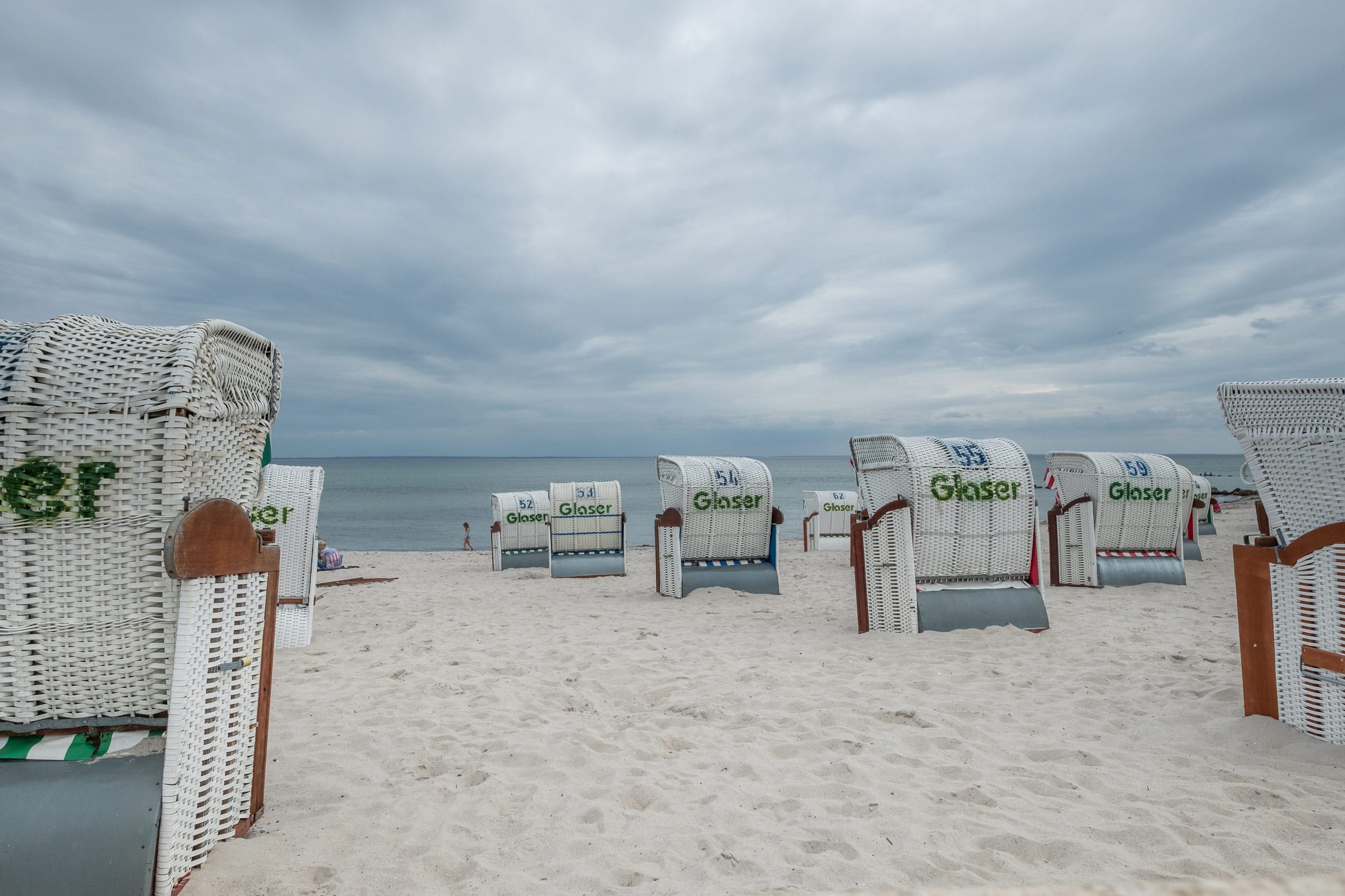 Grömitz im Winter an der Ostsee: Mehrere weiß-graue Strandkörbe stehen in Reihen auf einem feinsandigen Strand vor der ruhigen Ostsee unter einem bedeckten, grauen Himmel – nahe der Ostseeklinik Grömitz für Mutter-Kind-Kur und Vater-Kind-Kur.