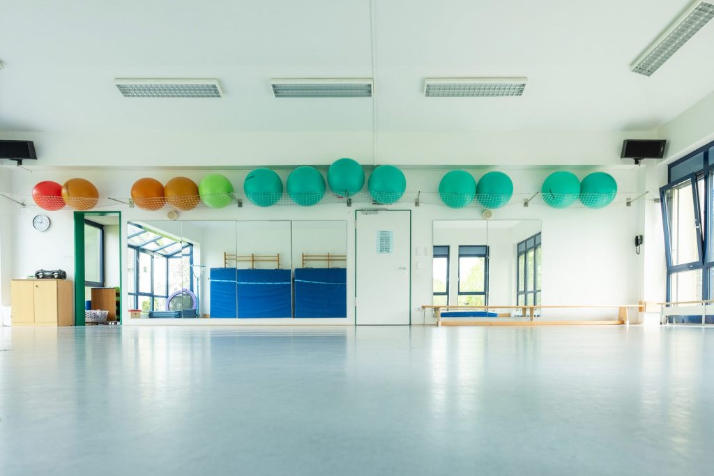 Ein heller, weitläufiger Gymnastikraum in der Ostseeklinik Grömitz mit hellem Boden und einer großen Spiegelwand. An einer Wandhalterung unter der Decke sind zahlreiche bunte Gymnastikbälle in Rot, Orange, Grün und Türkis aufgereiht. Große Fenster lassen viel Tageslicht in den freundlich gestalteten Therapieraum.