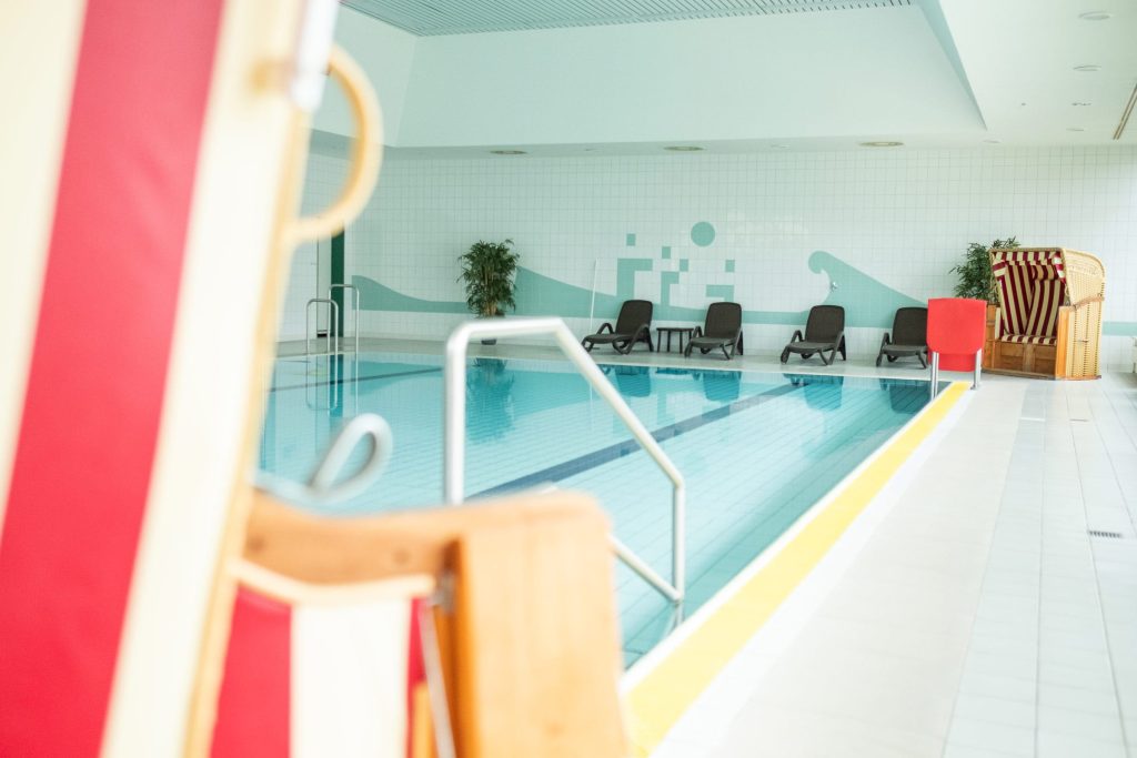 Heller Innenbereich des Schwimmbades der Ostseeklinik Grömitz mit blau schimmerndem Schwimmbecken, Liegestühlen am Beckenrand und typischem Ostsee-Strandkorb.