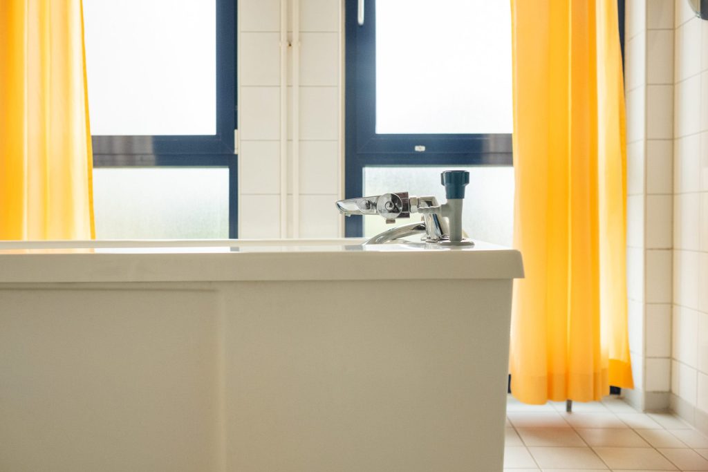 Seitenansicht einer weißen Therapiewanne in der Ostseeklinik Grömitz vor einem Fenster mit gelben Vorhängen und einer spezialisierten Armatur.