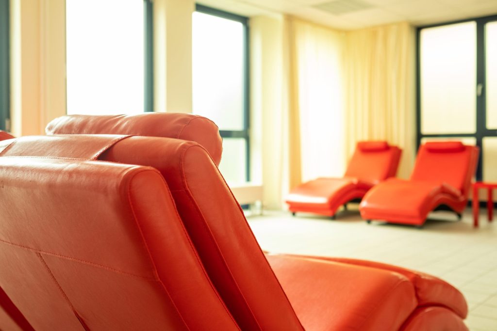 Nahaufnahme einer ergonomisch geformten, roten Leder-Relaxecke im Ruhebereich der Ostseeklinik Grömitz, im Hintergrund weitere Liegen vor hellen Fenstern.