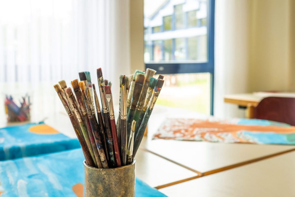 Nahaufnahme eines Bechers mit zahlreichen gebrauchten Pinseln in verschiedenen Größen vor einem hellen Kreativ- und Kunstraum mit gemalten Bildern.