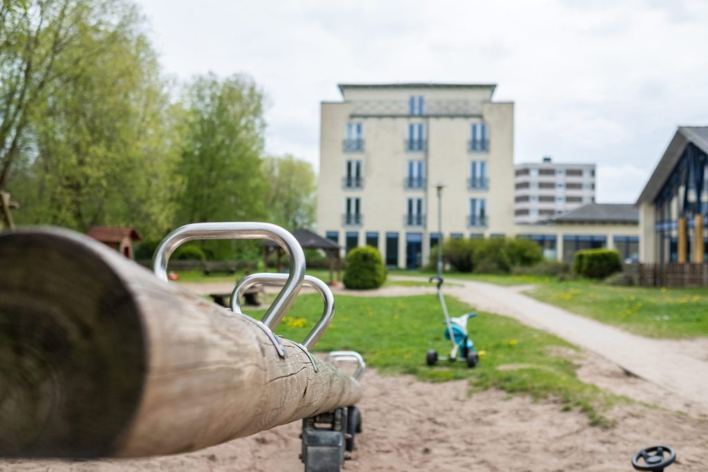 Blick über eine hölzerne Wippe auf dem Spielplatz zum Hauptgebäude der Ostseeklinik Grömitz.
