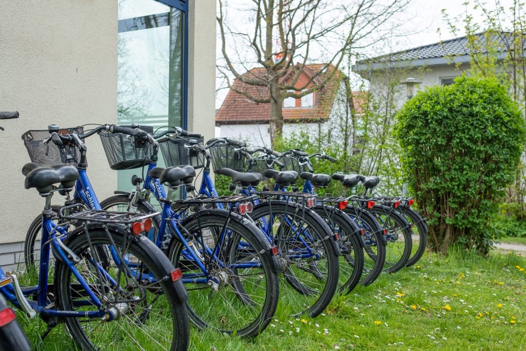 Eine Reihe baugleicher, blauer Fahrräder mit schwarzen Körben und Gepäckträgern steht ordentlich aufgereiht auf einer Rasenfläche vor einem Gebäude der Ostseeklinik Grömitz. Im Hintergrund sind Bäume und ein angrenzendes Wohnhaus mit Satteldach zu sehen.