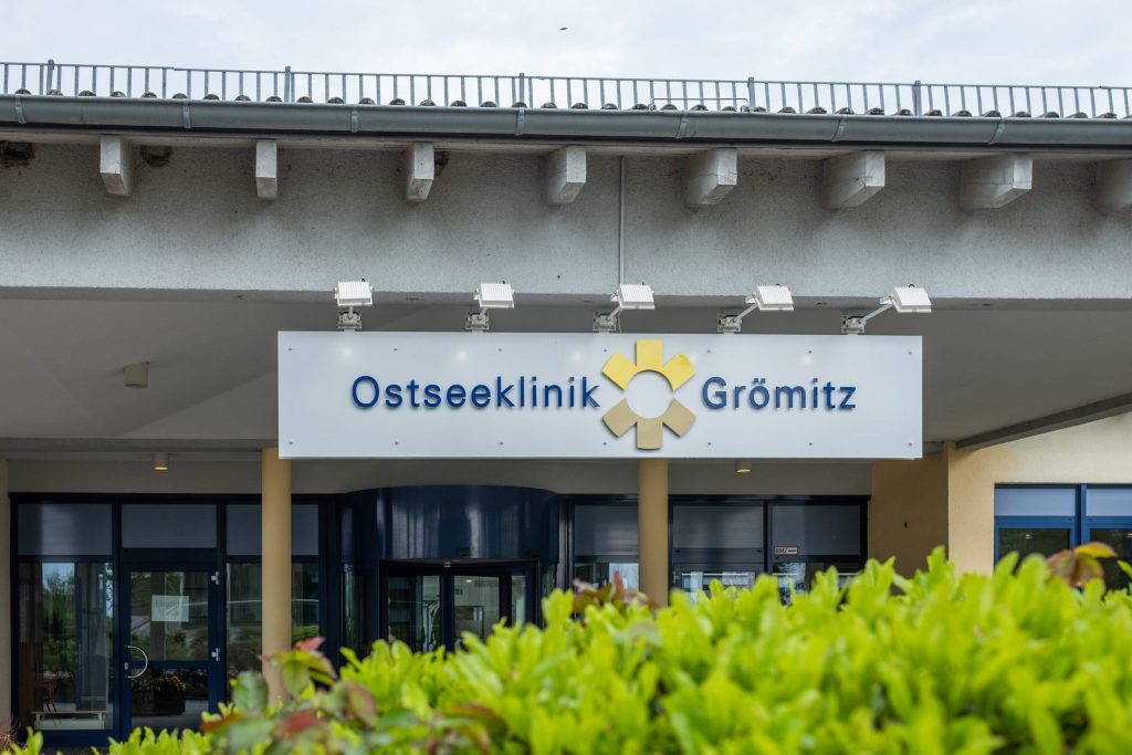Weißes Schild mit der blauen Aufschrift „Ostseeklinik Grömitz“ und einem gelben sonnenförmigen Logo über dem Haupteingang der Ostseeklinik Grömitz für Mutter-Kind-Kuren und Vater-Kinde-Kuren.