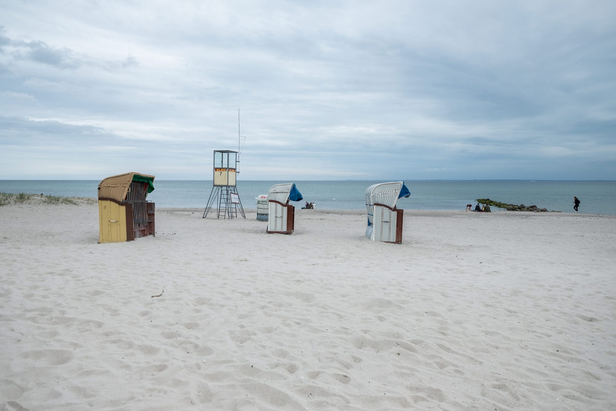 Lage und Umgebung Ostseeklinik Grömitz: Der Sandstrand direkt an der Rehaklinik für Mütter mit Kindern oder Väter mit Kindern: Zu sehen sind mehrere Strandkörbe, einem DLRG-Rettungsturm und der Blick auf die ruhige Ostsee unter leicht wolkigem Himmel.