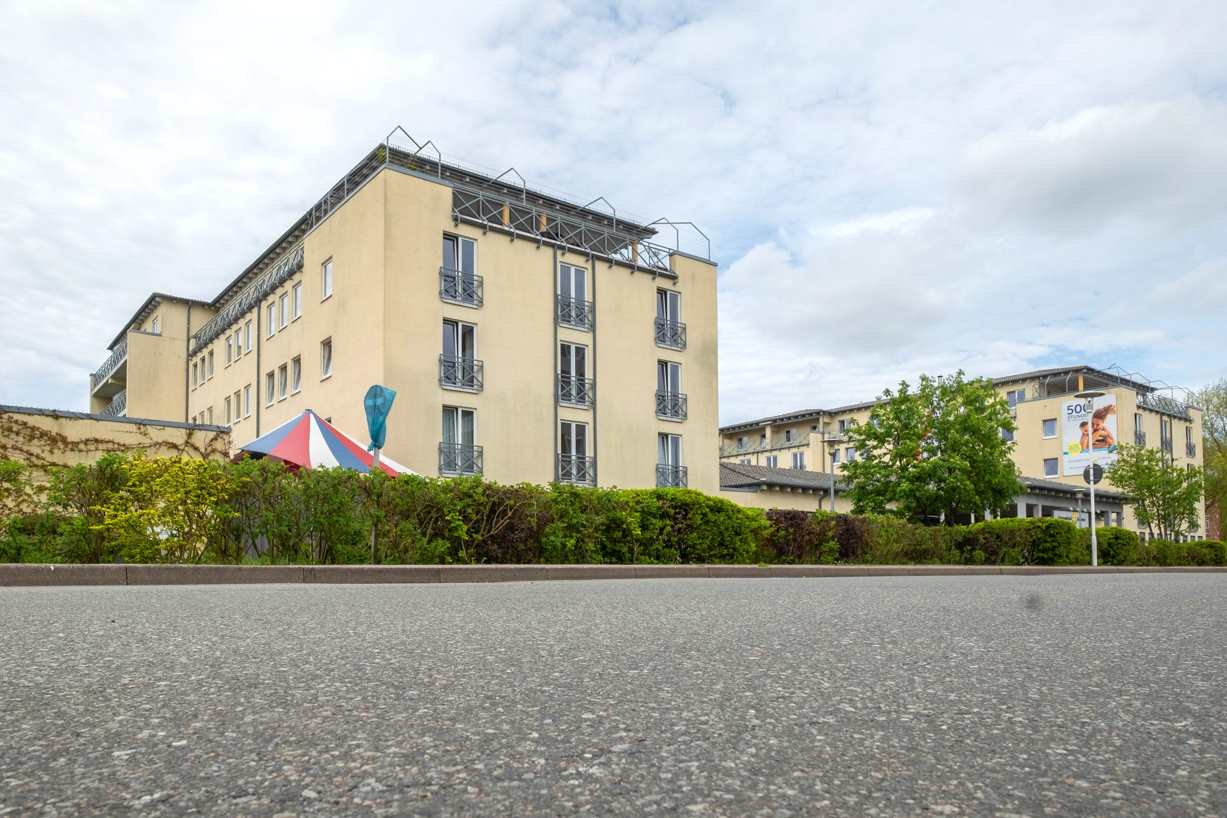 Außenansicht der Ostseeklinik Grömitz mit gelber Fassade, französischen Balkonen und einer gepflegten Hecke im Vordergrund unter leicht bewölktem Himmel