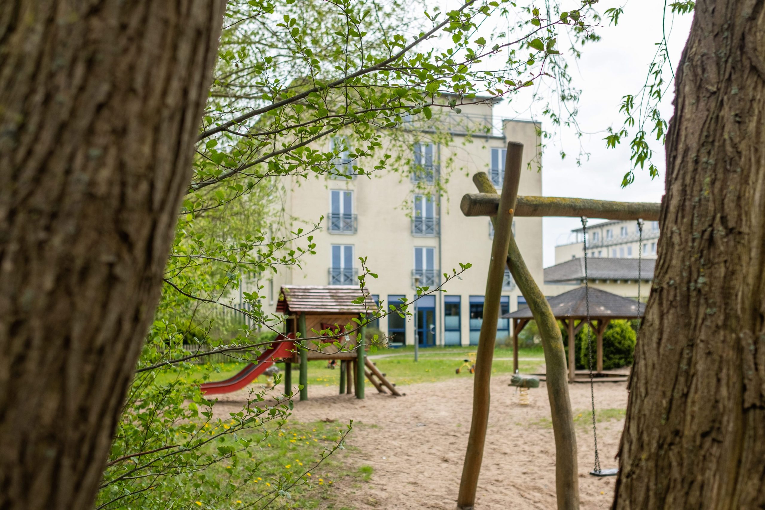 Kinderspielplatz der Mutter-Kind-Klinik in Grömitz: einladender Außenbereich mit Spielgeräten aus Holz und Blick auf das moderne Klinikgebäude, umrahmt von Bäumen.