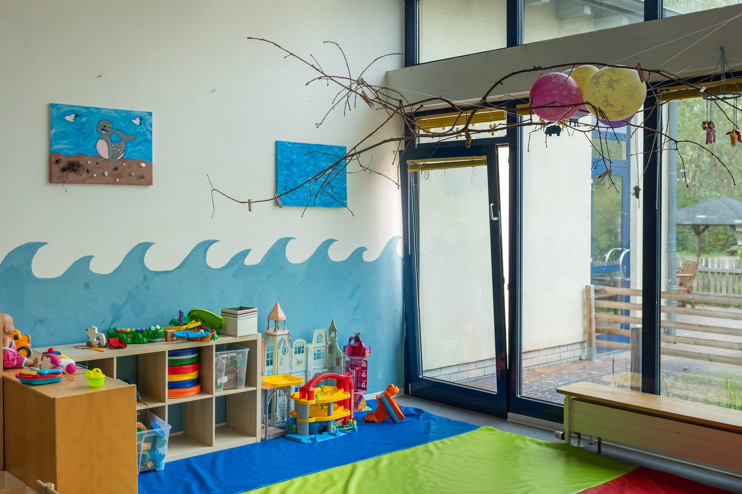 Ein helles, maritim gestaltetes Spielzimmer für Kinder in der Ostseeklinik Grömitz. Die Wand ist mit großen blauen Wellen bemalt, darüber ein blaues Leinwandbild und ein natürlicher Ast als Dekoration. Links davor Holzregale mit bunten Spielsachen, ein Spielzeug-Parkhaus und eine Spielburg auf einer blau-grünen Bodenmatte, rechts eine große Glasfront.