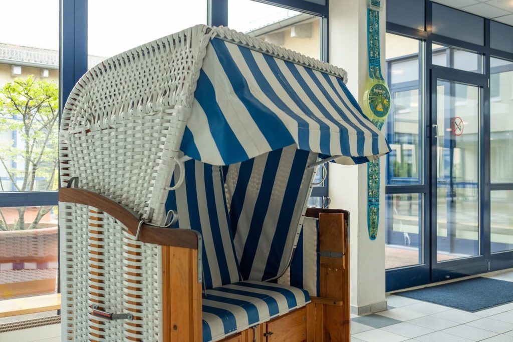 Ein klassischer, weiß geflochtener Strandkorb mit blau-weiß gestreifter Polsterung steht im hellen Eingangsbereich der Ostseeklinik Grömitz, direkt vor einer großen Fensterfront.