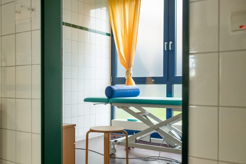 Ein heller Behandlungsraum in der Ostseeklinik Grömitz mit einer türkisfarbenen Behandlungsliege und einem Fenster mit gelben Vorhängen.