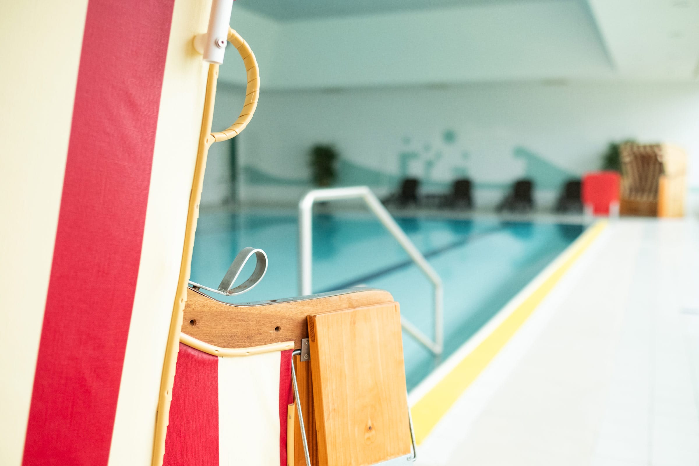 Nahaufnahme eines rot-weiß gestreiften Strandkorbs am Rande des hellen Hallenbads der Ostseeklinik Grömitz. Im unscharfen Hintergrund ist das tiefblaue Schwimmbecken mit Einstiegshilfe und Liegestühlen zu sehen.
