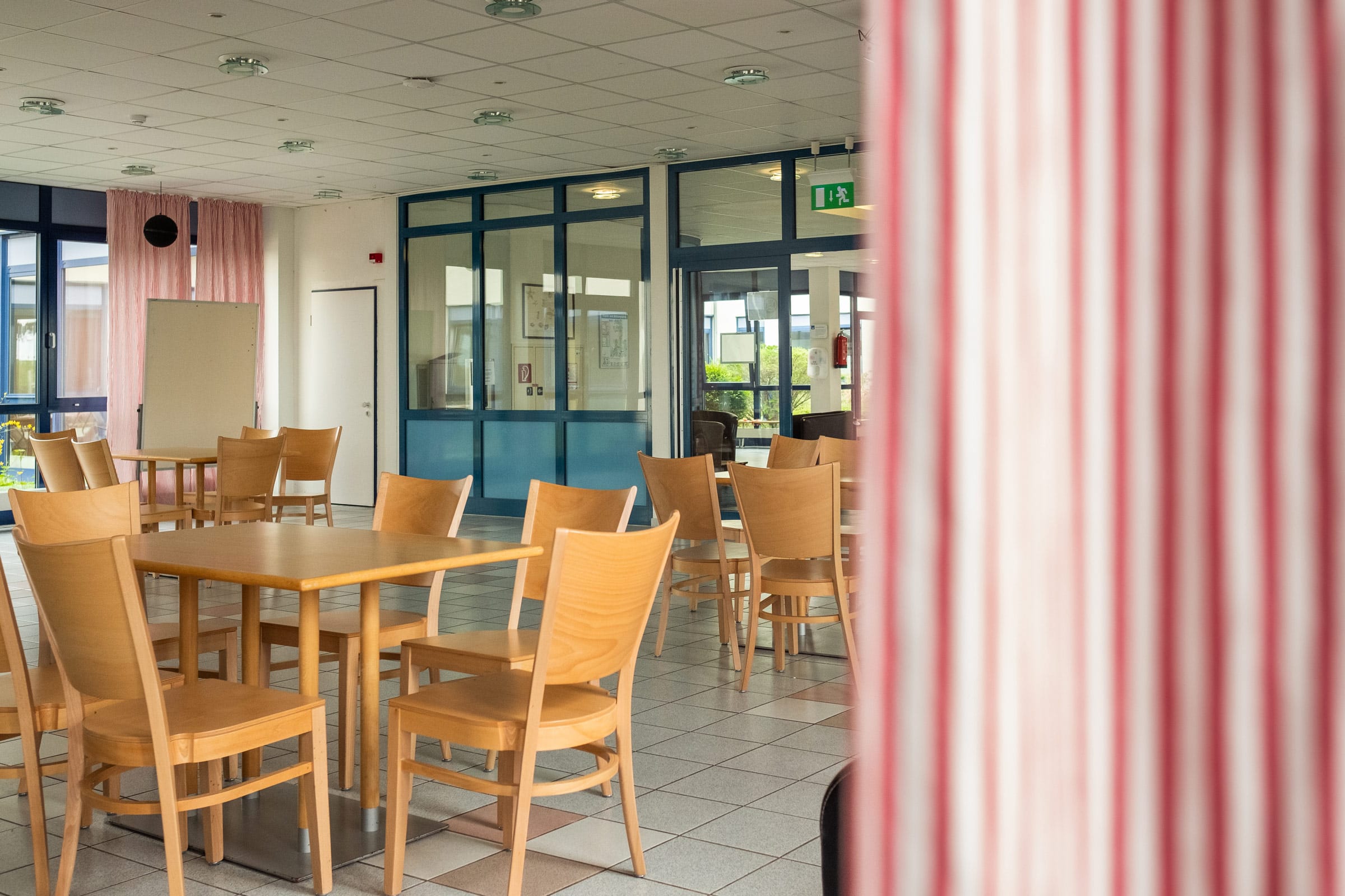 Helle Cafeteria mit mehreren Holztischen und passenden Holzstühlen auf einem hellen Fliesenboden. Im Hintergrund ist eine große Glasfront mit blauen Rahmen zu sehen.