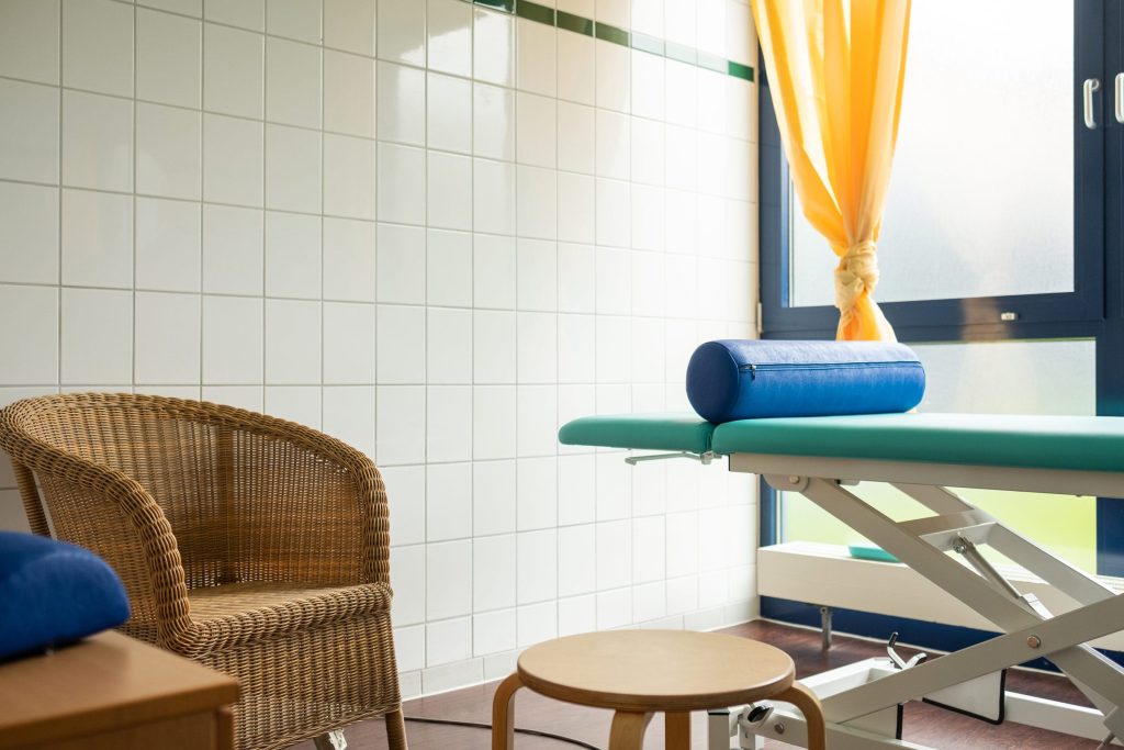 Ein heller Behandlungsraum in der Ostseeklinik Grömitz mit einer türkisfarbenen Therapieliege, einer blauen Lagerungsrolle, einem Korbsessel und einem Holzhocker vor einer weiß gefliesten Wand