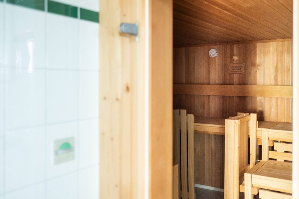 Blick durch eine geöffnete Holztür in eine Sauna der Ostseeklinik Grömitz. Der Innenraum ist komplett mit hellem Holz verkleidet und verfügt über mehrstufige Sitzbänke. An der Wand hängen ein Hygrometer und ein Hinweisschild. Links im Vordergrund ist ein Teil der weiß gefliesten Wand des Vorraums mit grünen Akzentfliesen zu sehen.