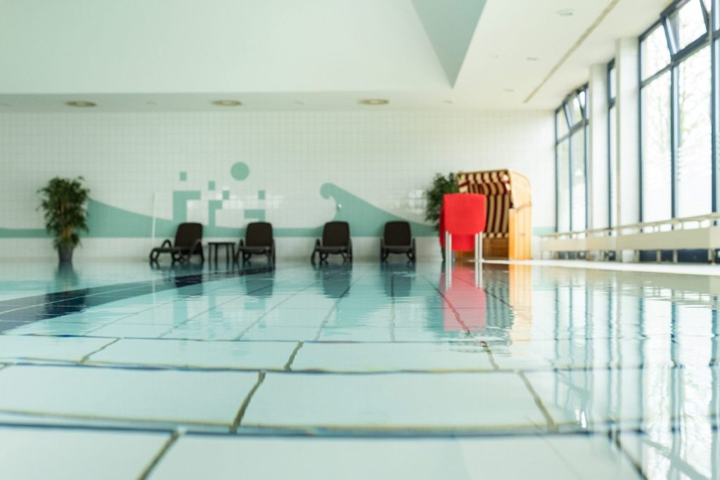 Hell gefliestes Schwimmbad der Ostseeklinik Grömitz. Unten Spiegelungen im Wasser, im Hintergrund eine große Topfpflanze, dunkle Liegen und ein Strandkorb, rechts eine große Fensterfront.