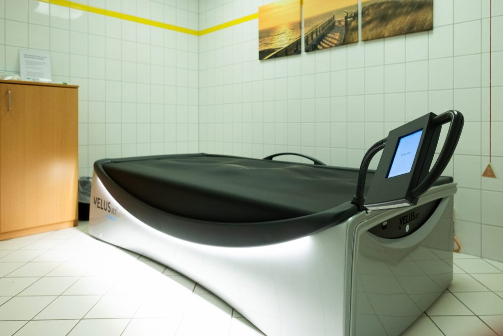 Ein heller, gefliester Behandlungsraum mit einer modernen Hydrojet-Massage-Liege im Vordergrund. Die Liege ist silberweiß mit einer schwarzen Auflage; an der Wand dahinter hängen drei Leinwandbilder, die eine Dünenlandschaft bei Sonnenuntergang zeigen.