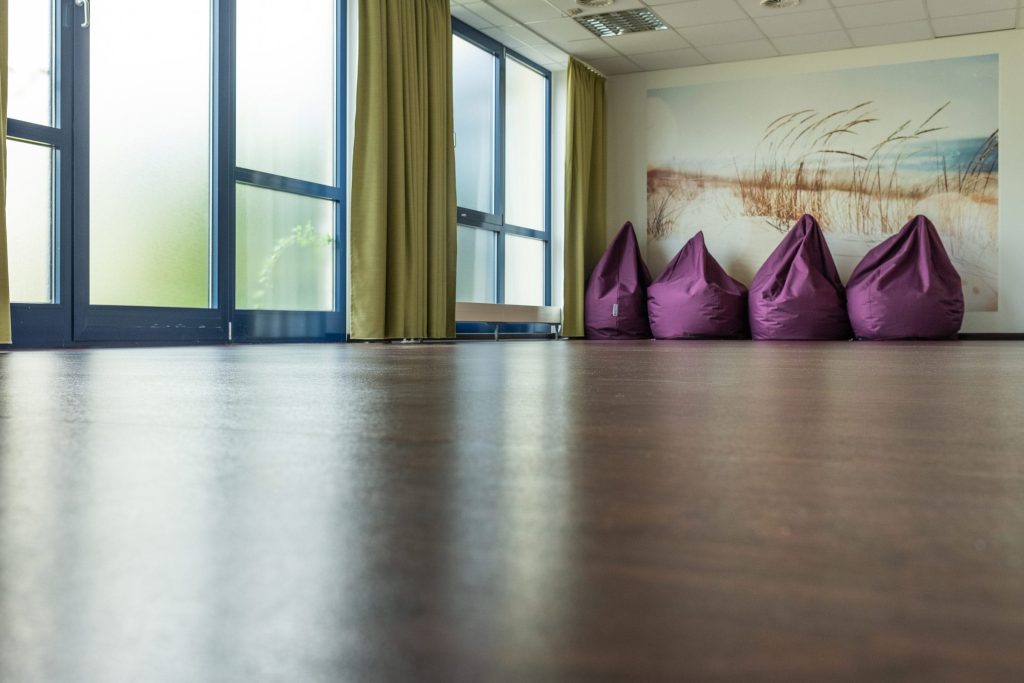 Heller, moderner Therapie- und Entspannungsraum in der Ostseeklinik Grömitz mit dunklem Bodenbelag, großen Fensterfronten und olivgrünen Vorhängen. An der Stirnseite befinden sich vier lilafarbene Sitzsäcke vor einer Wandtapete, die eine beruhigende Dünenlandschaft an der Ostsee zeigt.