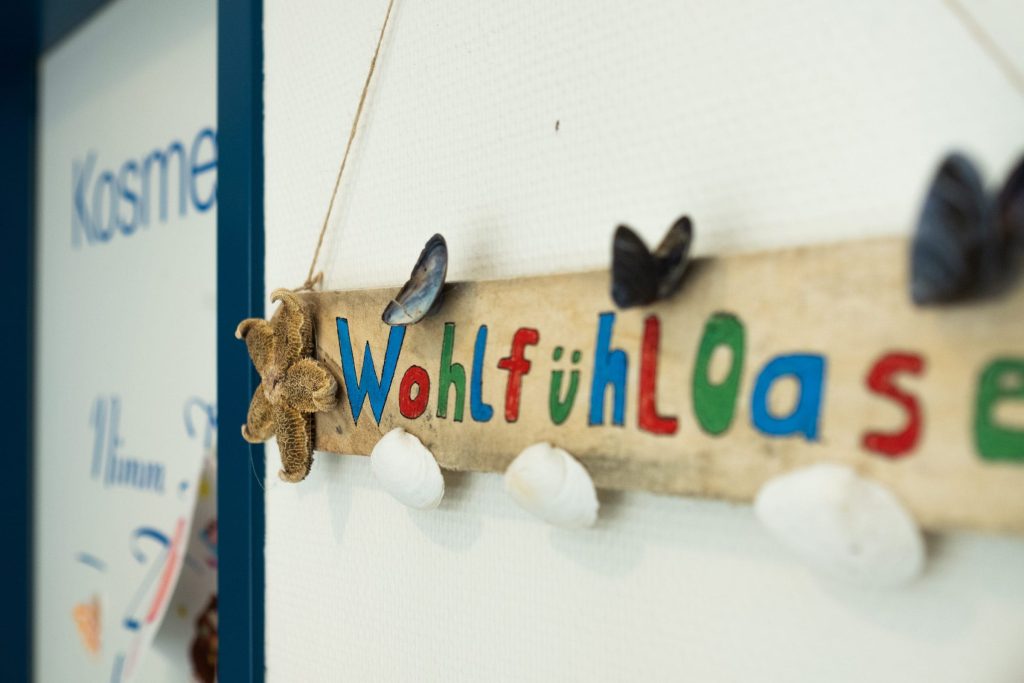 Ein dekoratives Holzschild an einer weißen Wand in der Ostseeklinik Grömitz. Auf dem rustikalen Treibholz steht in bunten, verspielten Buchstaben das Wort „Wohlfühloase“. Das Schild ist maritim mit Muscheln, kleinen blauen Schmetterlingsfiguren und einem Seestern verziert. Im unscharfen Hintergrund ist ein Türrahmen mit der Aufschrift „Kosmetik“ zu erkennen.