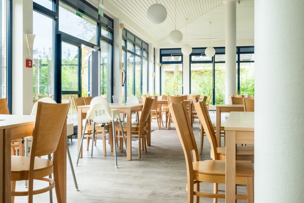 Ein heller, lichtdurchfluteter Speisesaal mit großen Glasfronten, die den Blick ins Grüne freigeben. Im Raum stehen mehrere helle Holztische mit passenden Stühlen sowie weiße Kinderhochstühle; schlichte, kugelförmige Pendelleuchten hängen von der Decke.