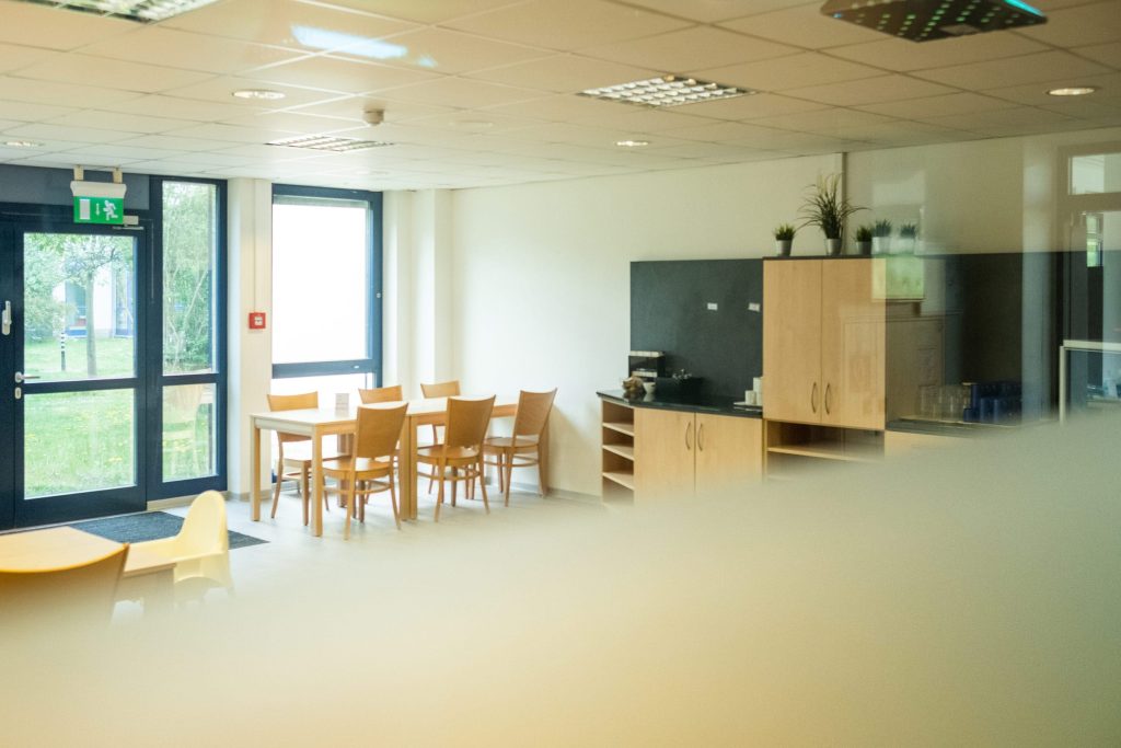 Helle, modern gestalteter Gemeinschaftsraum. Im Zentrum ein Holztisch mit passenden Stühlen vor einem großen Fenster, das den Blick ins Grüne freigibt. Rechts eine Küchenzeile mit hellen Holzfronten und einer dunklen Rückwand. Ein Notausgang mit grüner Kennzeichnung führt direkt nach draußen.