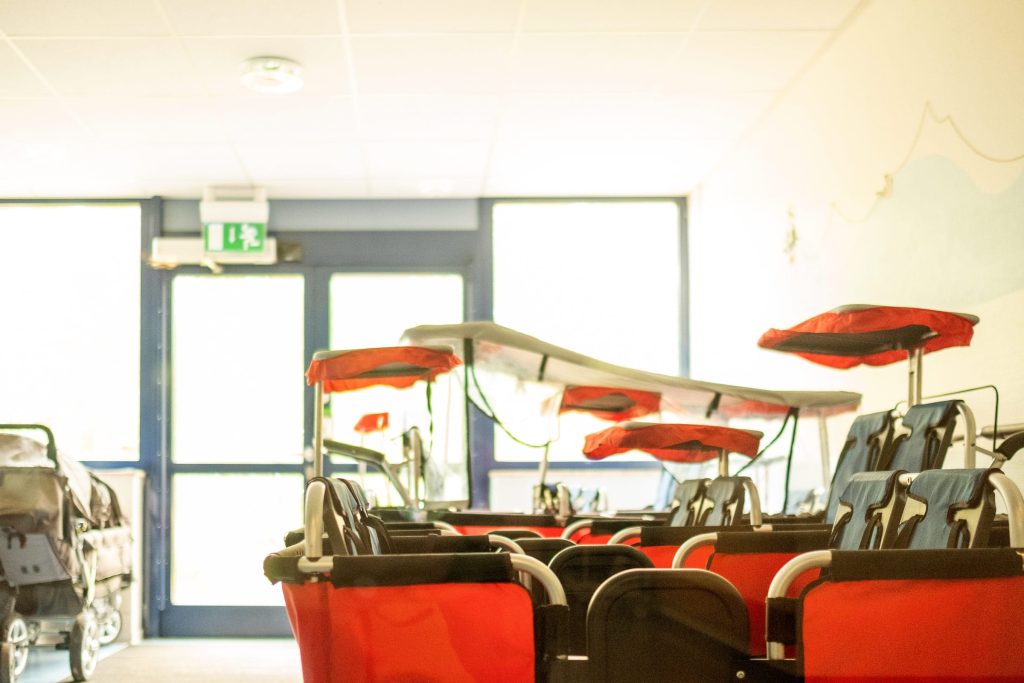 Mehrere rote Mehrsitzer-Kinderwagen (Krippenwagen) mit roten Sonnendächern stehen ordentlich aufgereiht in einem hellen Innenraum der Ostseeklinik Grömitz vor einer Glastür mit Notausgangsschild.