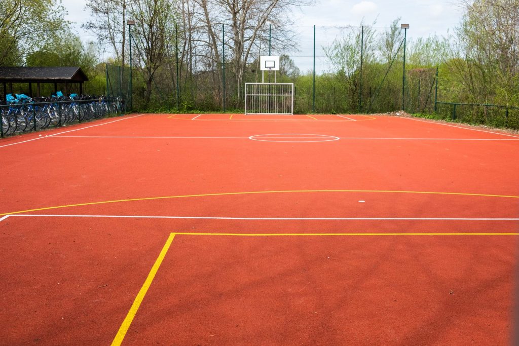 Ein moderner Mehrzweck-Sportplatz im Außenbereich der Ostseeklinik Grömitz mit rotem Bodenbelag, Basketballkorb und integriertem Fußballtor. Im Hintergrund sind grüne Bäume und ein Fahrradunterstand zu sehen.