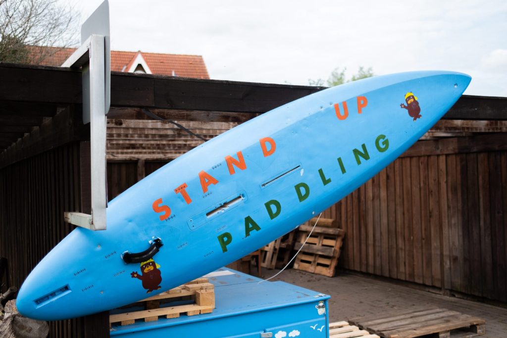 Ein blaues Stand-Up-Paddling-Board lehnt diagonal an einer Holzhütte im Außenbereich der Ostseeklinik Grömitz. Das Board trägt die Aufschrift „STAND UP PADDLING“ in bunten Buchstaben und ist mit zwei Illustrationen des Maskottchens Grömel mit gelber Kappe verziert.