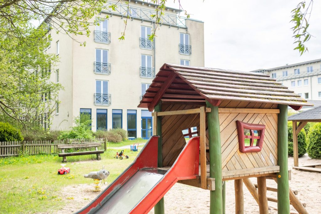Ein Spielplatz im Außenbereich der Ostseeklinik Grömitz mit einem hölzernen Spielhaus inklusive Rutsche, einer Wippe in Robbenform und einer hölzernen Sitzbank vor einem mehrstöckigen Klinikgebäude mit Balkonen.