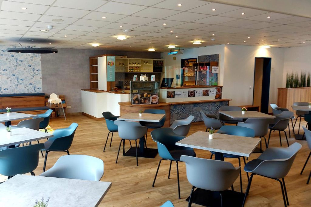 Modern eingerichtete Cafeteria der Ostseeklinik Grömitz mit hellem Holzfußboden und grauen Tischen. Im Vordergrund zahlreiche graue und blaue Schalensitze an quadratischen Tischen. Im Hintergrund befindet ein heller Verkaufstresen mit einer Glasvitrine für Gebäck und einem hölzernen Regalsystem an der Rückwand. Die Decke ist mit vielen kleinen Einbaustrahlern versehen, die den Raum warm beleuchten. Links im Bild eine Wand mit blau gemusterter Tapete und einer hölzernen Sitzbank.