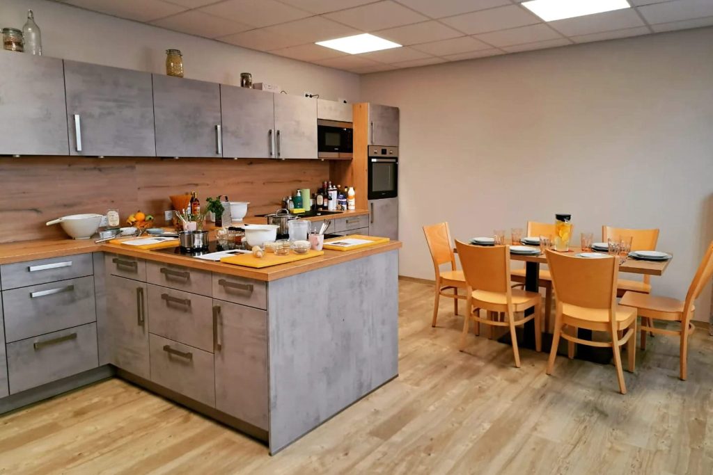 Moderne, hell beleuchtete Lernküche mit einer Kochinsel in Beton-Optik und Holz-Arbeitsflächen. Auf der Insel und der hinteren Küchenzeile stehen verschiedene Kochutensilien und Zutaten bereit. Im Hintergrund ist ein angrenzender Essbereich mit einem gedeckten Holztisch und sechs passenden Stühlen auf hellem Laminatboden zu sehen.