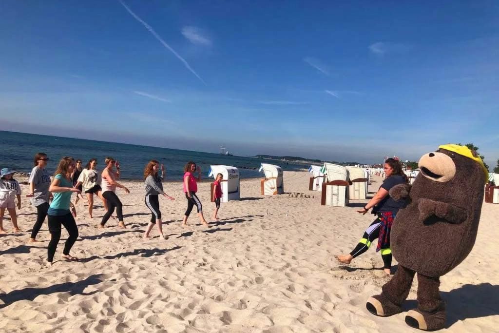 Eine Gruppe von Müttern und Kindern bei einem Fitnesskurs am Sandstrand an der Ostseeklinik Grömitz. Rechts im Vordergrund macht Seebär Grömel als lebensgroßes, braunes Maskottchen mit. Im Hintergrund sind das blaue Meer, ein strahlend blauer Himmel und mehrere typische weiße Strandkörbe zu sehen.
