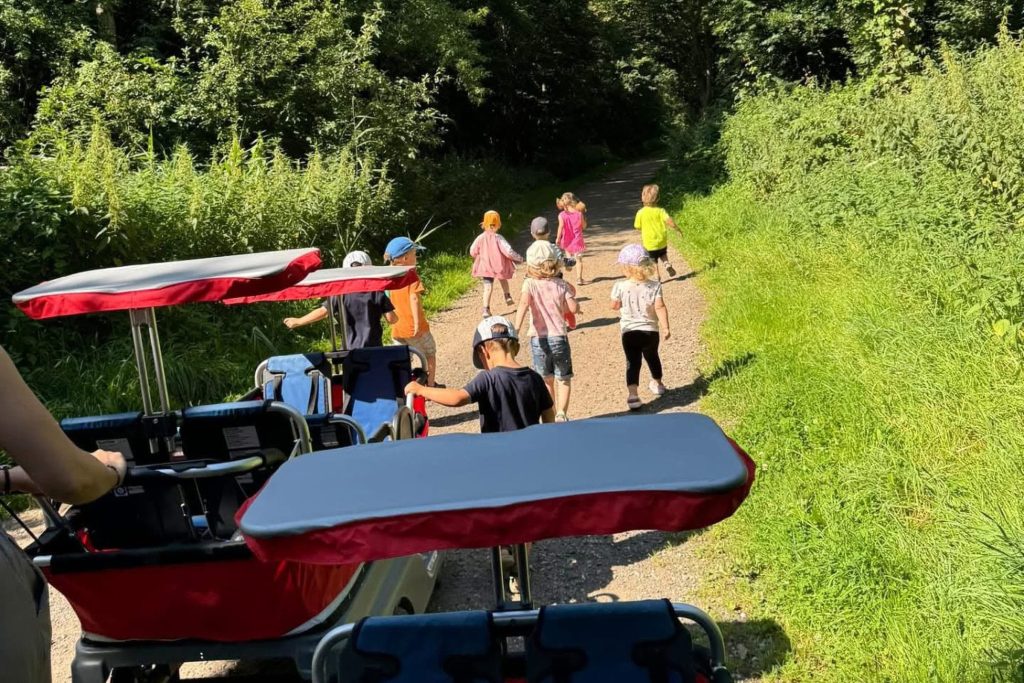 Eine Gruppe von Kindern der Ostseeklinik Grömitz bei einem Ausflug auf einem sonnigen Waldweg, begleitet von zwei großen, roten Krippenwagen mit Sonnendächern.
