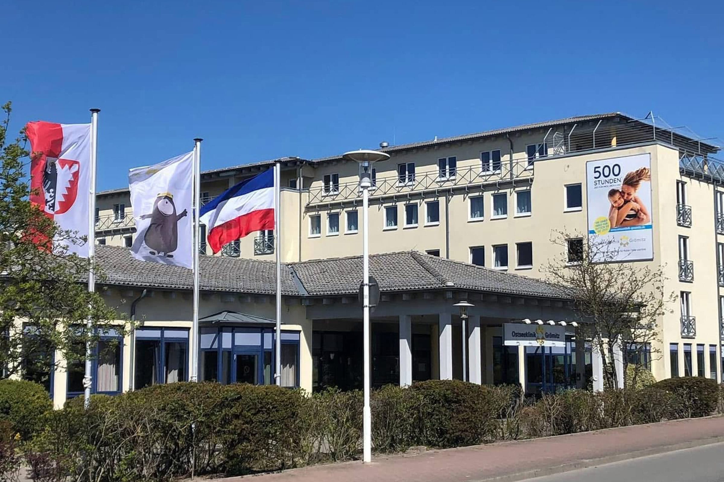 Außenansicht der Ostseeklinik Grömitz bei sonnigem Wetter. Im Vordergrund links wehen drei Flaggen (Schleswig-Holstein, das Maskottchen Grömitz und ein Wappen) vor einem modernen, mehrstöckigen Klinikgebäude mit gelber Fassade. An der rechten Außenwand hängt ein großes Plakat mit der Aufschrift „500 Stunden“, das eine Mutter mit ihrem Kind zeigt.