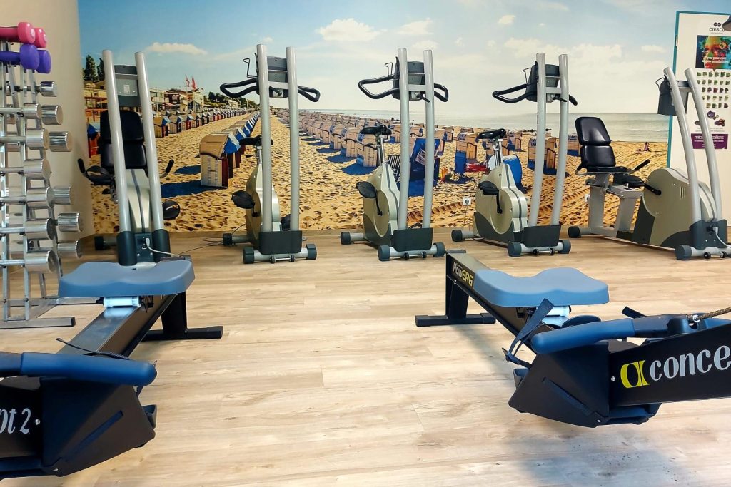 Heller Trainingsraum in der Ostseeklinik Grömitz mit modernen Fitnessgeräten vor einer großflächigen Wandtapete, die einen Ostseestrand mit zahlreichen Strandkörben zeigt. Im Vordergrund stehen zwei Rudergeräte, dahinter sind mehrere Ergometer und ein Hantelständer auf hellem Holzboden angeordnet.