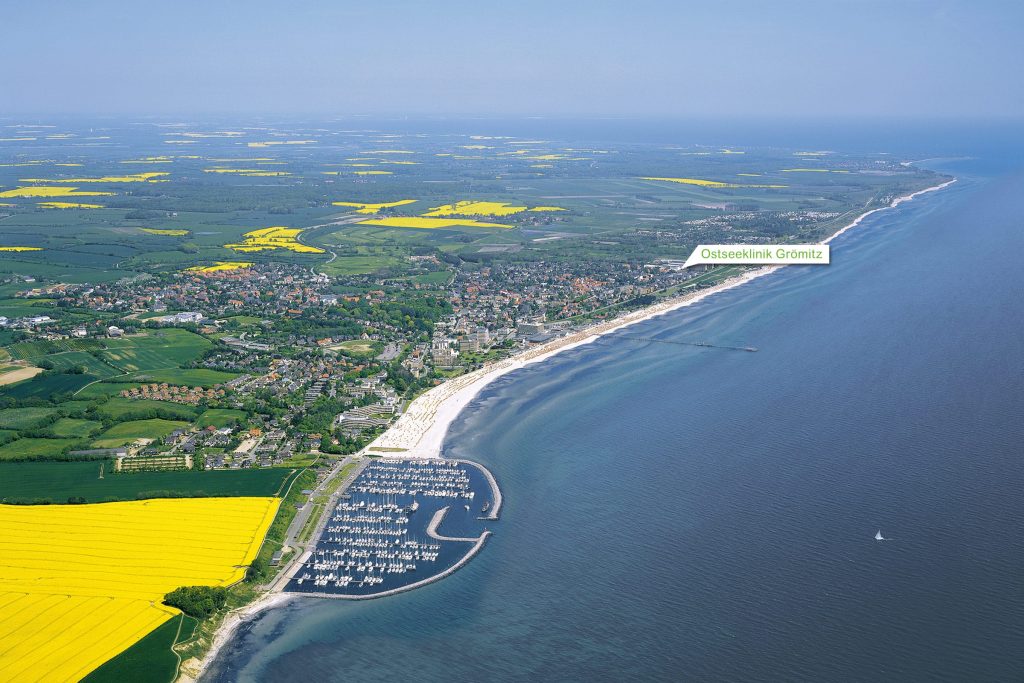 Luftaufnahme der Ostseeküste bei Grömitz: Yachthafen mit Segelbooten, gelbe Rapsfelder, weißer Sandstrand und blaues Meer. Eine Textmarkierung weist auf die „Ostseeklinik Grömitz" hin.