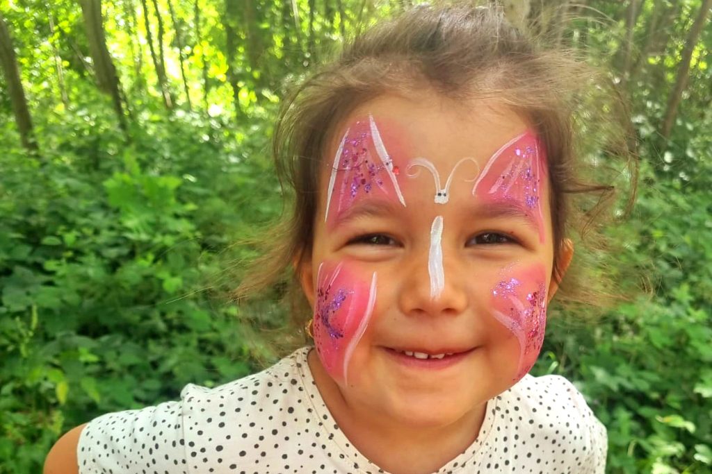 Ein lächelndes junges Mädchen mit buntem Schmetterlings-Kinderschminken im Gesicht. Die Flügel sind in Rosa und Weiß mit glitzernden Akzenten gestaltet. Im Hintergrund ist eine grüne, bewaldete Naturkulisse der Ostseeklinik Grömitz zu sehen.