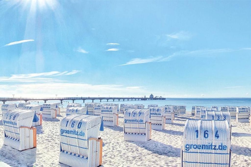 Zahlreiche weiß-blaue Strandkörbe im feinen Sand am Grömitzer Ostseestrand. Im Hintergrund die Seebrücke Grömitz unter strahlend blauen Himmel bei Sonnenschein. Auf den Strandkörben sind der Schriftzug "groemitz.de" sowie individuelle Nummern wie 160 und 164 zu sehen.