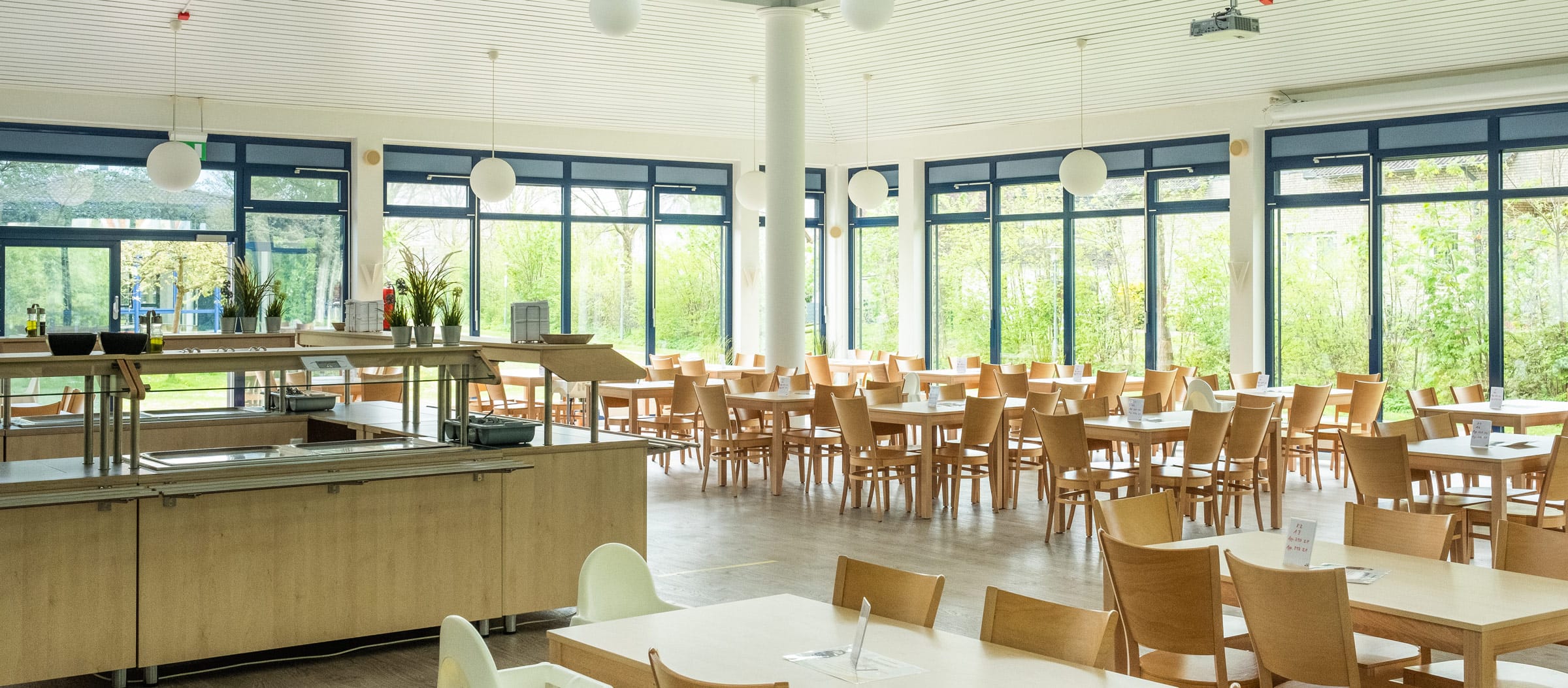 Lichtdurchfluteter Speisesaal der Ostseeklinik Grömitz mit großen Panoramafenstern und Blick ins Grüne. Zahlreiche Holztische und Stühle, links ein Buffetbereich mit heller Holzverkleidung, moderne Pendelleuchten an der Decke. 