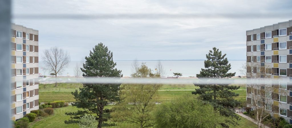 Blick aus einem Fenster der Ostseeklinik Grömitz auf die gepflegten Grünanlagen, hohe Nadelbäume und zwei mehrstöckige Klinikgebäude mit farbigen Balkonakzenten, im Hintergrund der Ostseestrand und das Meer unter bewölktem Himmel.