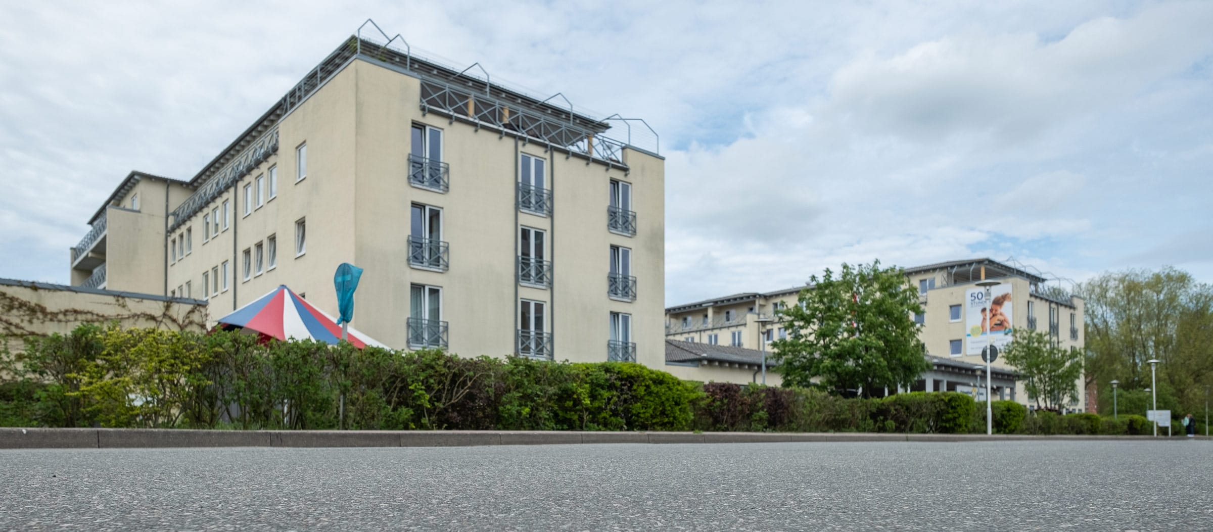 Die Ostseeklinik Grömitz: Ansicht der Patientenappartements und des Außengeländes der Mutter/Vater-Kind-Vorsorge- und Rehabilitationsklinik: mehrstöckige, helle Klinikgebäude der Kur- und Rehaklinik hinter einer grünen Hecke und blauem Himmel mit weißen Wolken.
