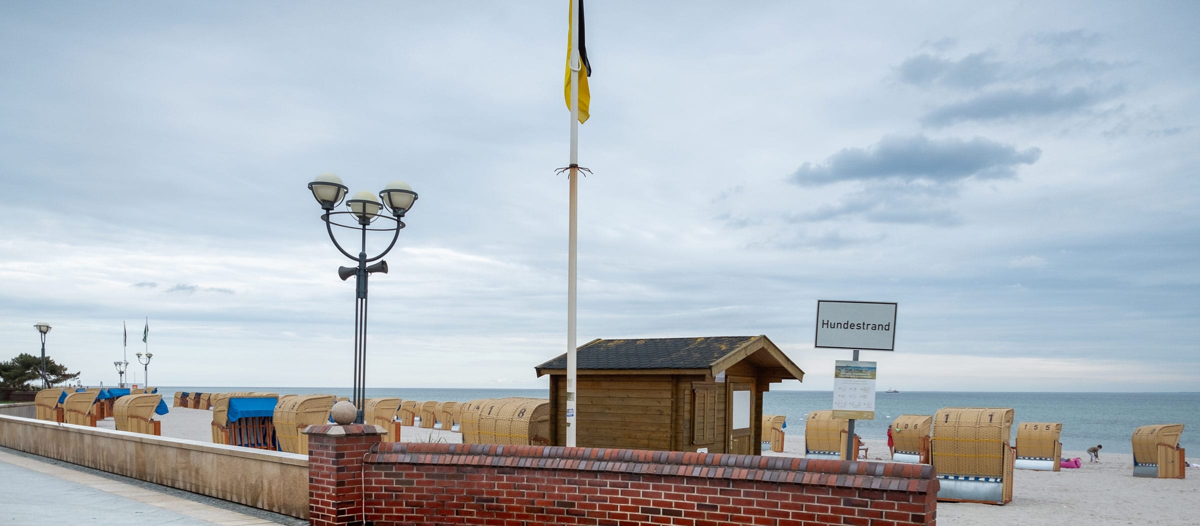 Blick auf den weitläufigen Strand an der Ostsee in Grömitz. Im Vordergrund eine Backsteinmauer und die Strandpromenade mit einer nostalgischen Straßenlaterne. Auf dem Sandstrand stehen viele Strandkörbe, eine kleine Holzhütte sowie ein Schild mit der Aufschrift „Hundestrand“. Im Hintergrund erstreckt sich die Ostsee unter einem wolkenverhangenen Himmel.